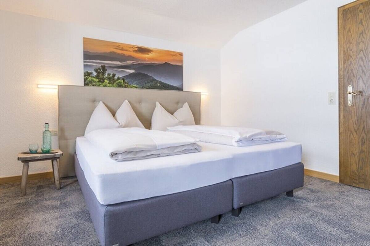 A neatly made bed with a headboard, a small bedside table with a bottle and glass, and a painting of a landscape on the wall.