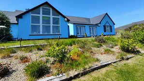 Uma casa azul de dois andares, com janelas amplas e telhado inclinado, cercada por um jardim bem cuidado com diversas plantas e um caminho de cascalho.