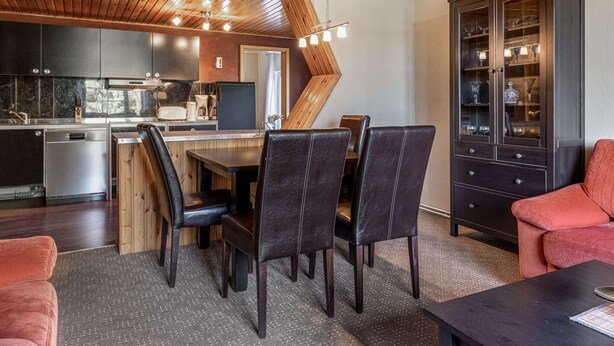Une cuisine moderne avec des meubles de couleur sombre, un plafond en bois et un coin repas doté de chaises en cuir et d’un canapé rouge.