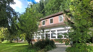 A two-story building with a red-tiled roof, surrounded by lush greenery and a gravel path leading to the entrance.