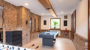 A room with a pool table, foosball table, brick walls, and a window with a plant. - Holiday Home With Sauna in the Eifel National Park (Schleiden)