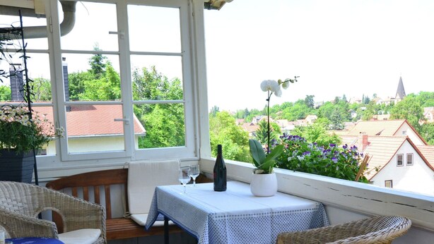 Uma varanda com móveis de vime, uma mesa com toalha azul e um vaso com planta.