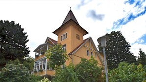 Um edifício amarelo com uma torre, rodeado por vegetação e uma parede de pedra.