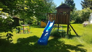 Ein Spielplatz mit einer blauen Rutsche, einem hölzernen Spielhäuschen und einem kleinen Tisch mit Stühlen.