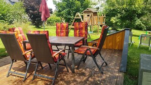 Eine Außenpatio mit Holzterrasse, Korbstühle mit roten und gelben Kissen, einem Metalltisch und einem hölzernen Spielhaus im Hintergrund.