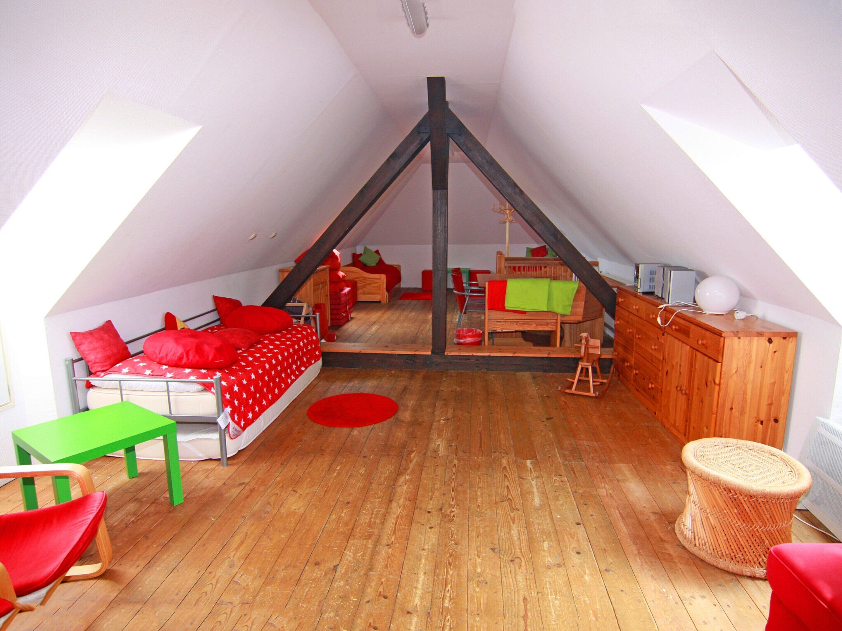 A cozy attic bedroom with a sloped ceiling, a bed with red bedding, a green table, and a wooden dresser.