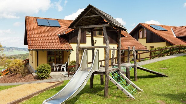 Un terrain de jeux avec un toboggan et des structures d’escalade, situé devant un bâtiment doté d’un toit en pente et de panneaux solaires.