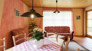 Un salon chaleureux avec un plafond en bois, un canapé rouge, une table à manger en bois recouverte d’une nappe à carreaux et deux lampes suspendues.