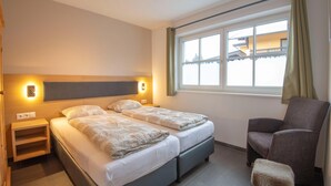 Hotellihuone, jossa on kaksi sänkyä, tuoli, pieni jakkara, yöpöytä ja ikkuna, jossa on verhot.