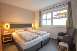 Quarto de hotel com duas camas, uma cadeira, um banquinho, uma mesa de cabeceira e uma janela com cortinas.