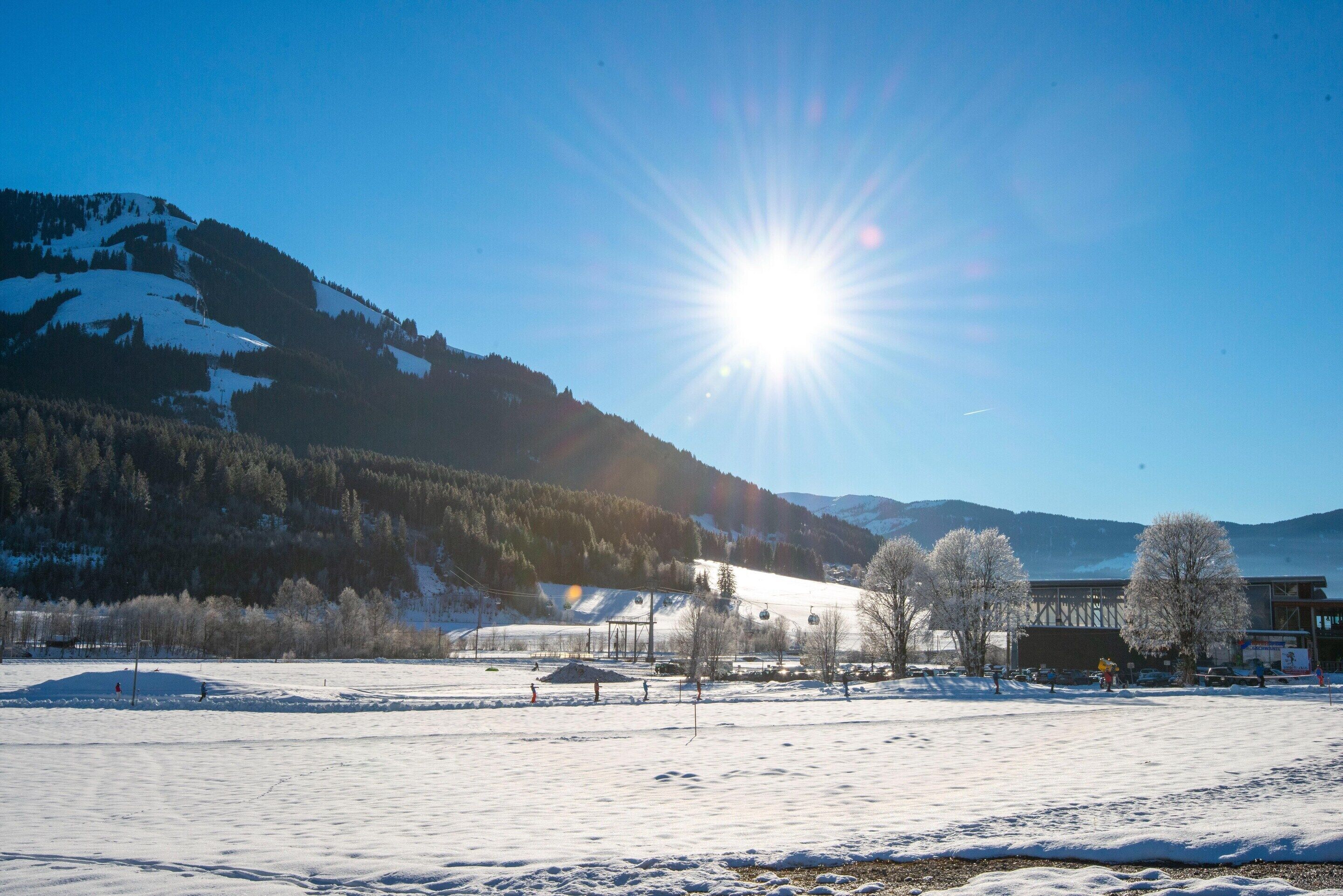 A snow-covered landscape with a clear sky, mountains, and a building with a ski lift in the background.