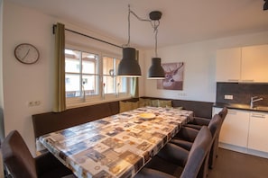 Uma sala de jantar com uma mesa comprida, cadeiras, luminárias pendentes, um relógio e uma janela com cortinas.