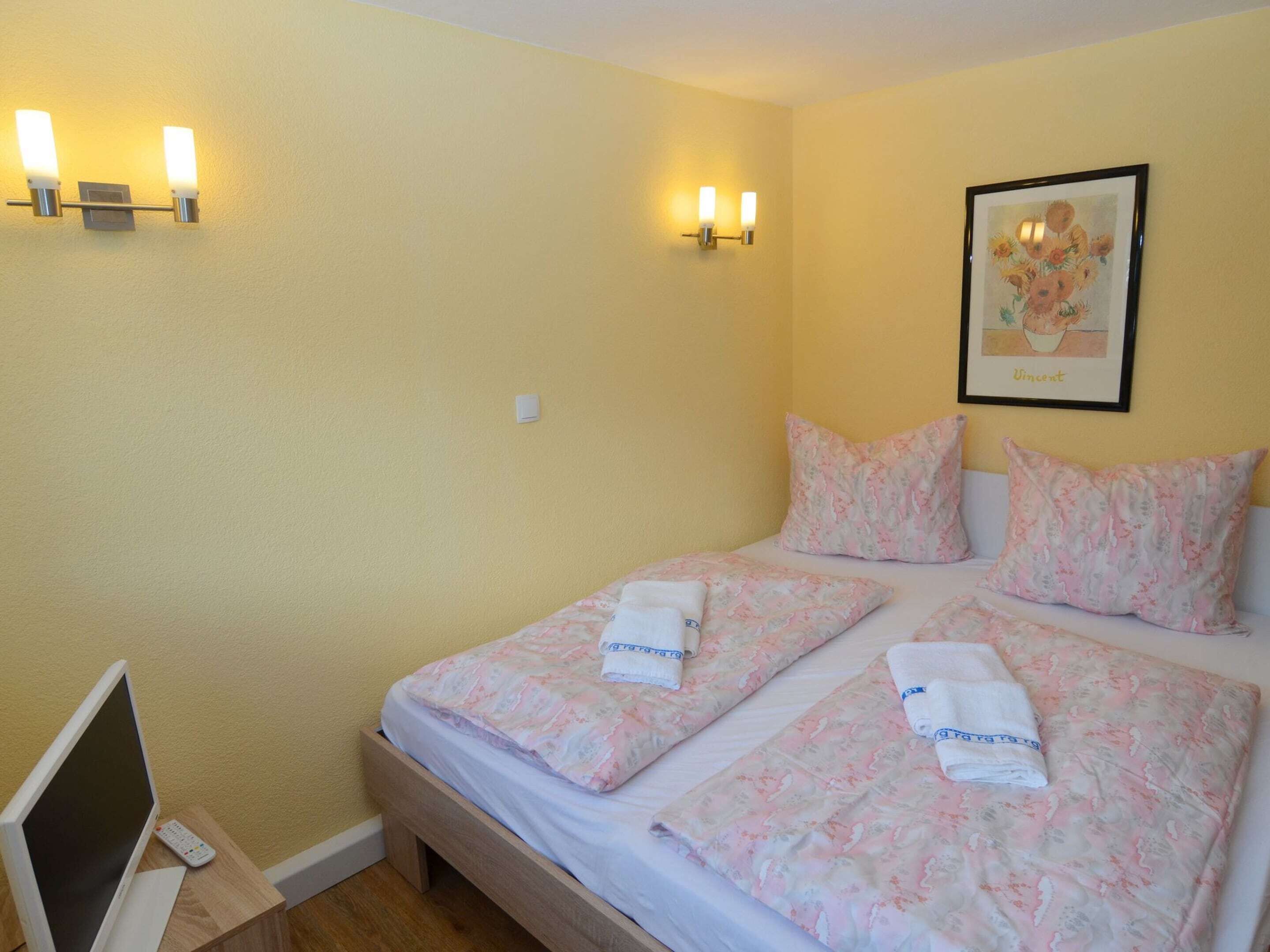 A hotel room with two beds, each with floral bedding and towels. There are wall-mounted lights and a framed picture on the wall.