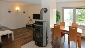 Ein modernes Wohnzimmer mit Kaminofen, einem Essbereich mit Holzsesseln, einem weißen Sofa und einem Fernseher an der Wand.