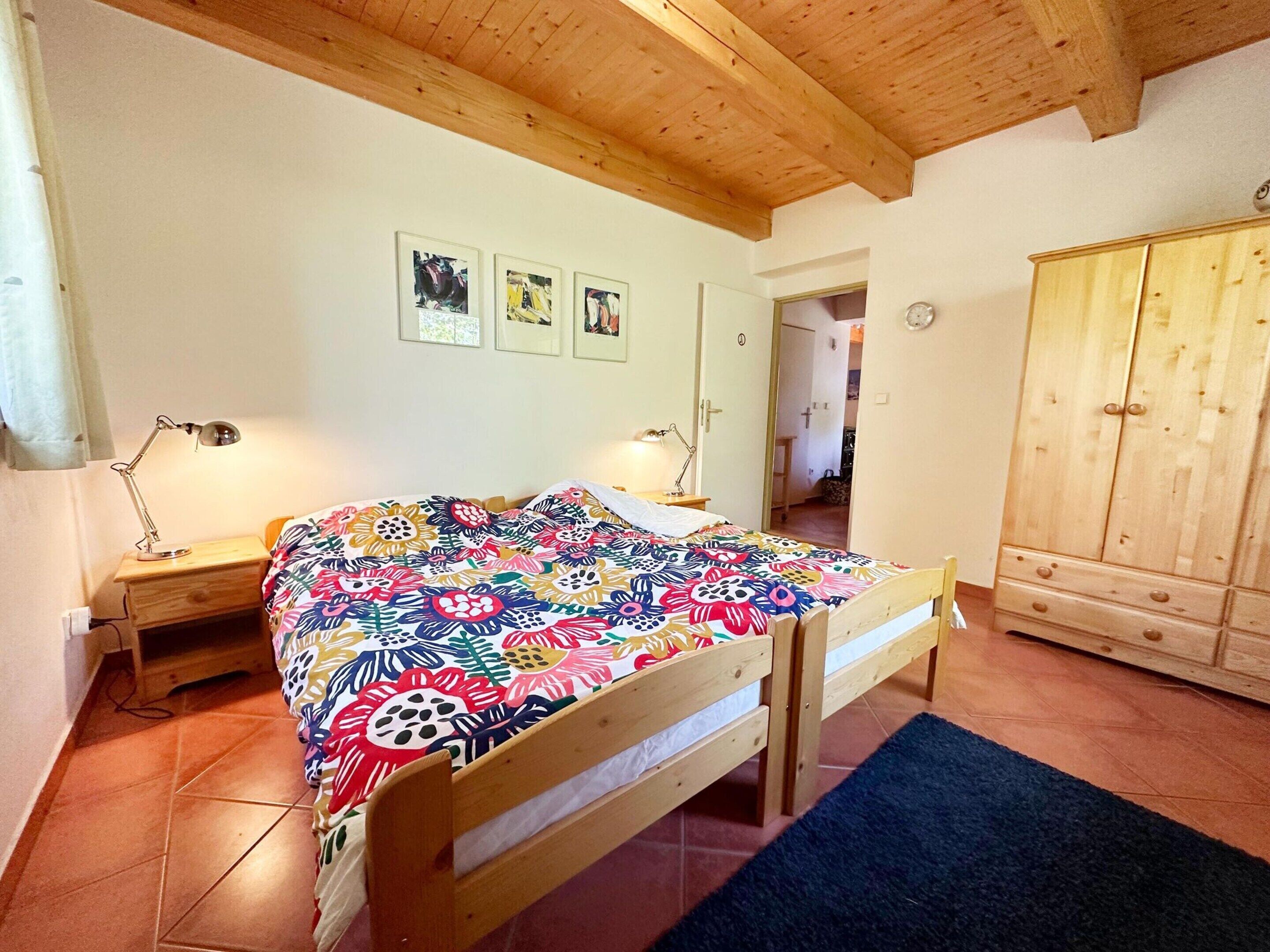 A bedroom with a wooden bed, two bedside tables with lamps, a colorful floral bedspread, and a wardrobe.