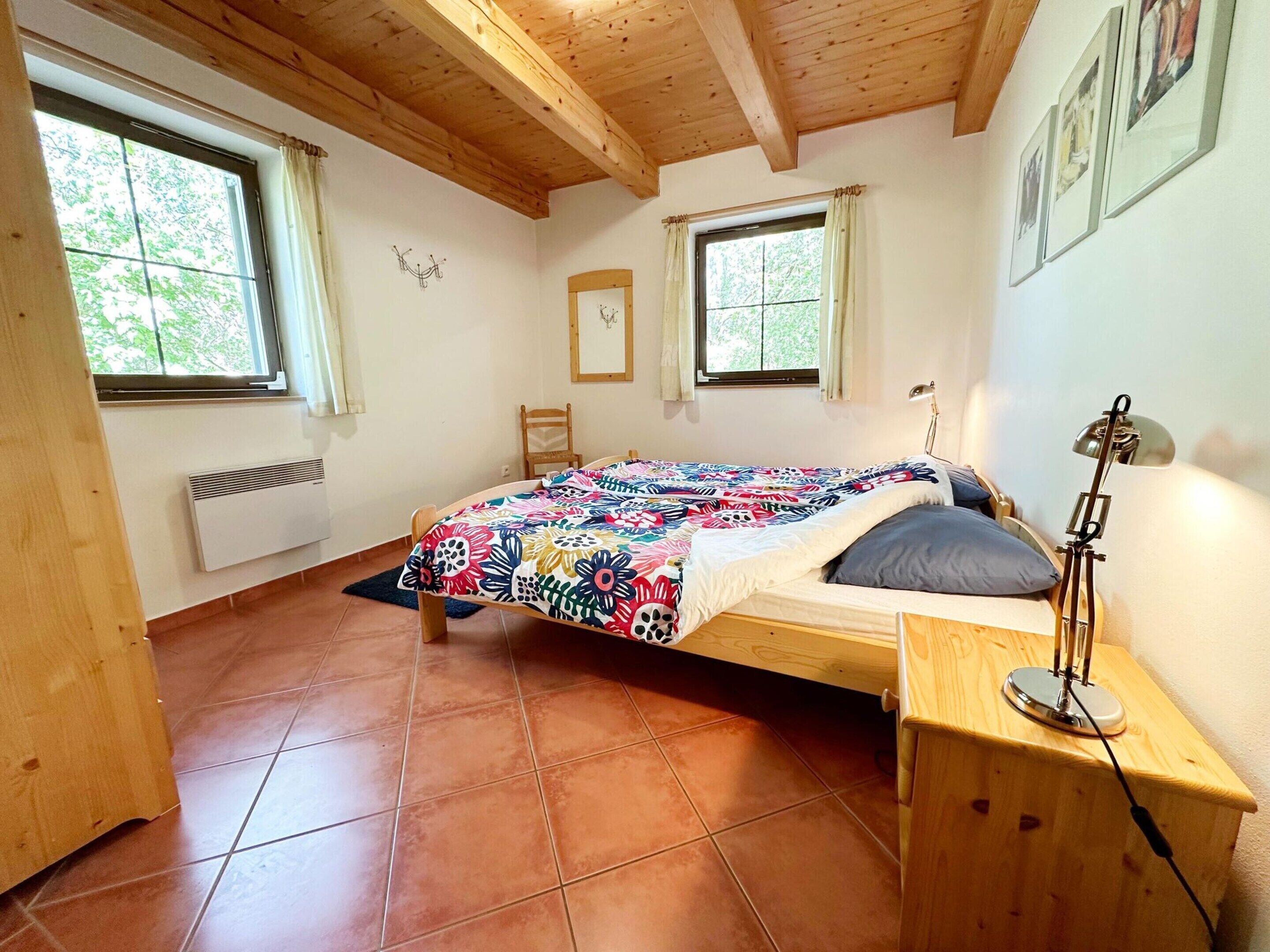 A bedroom with a wooden bed, a desk, and a chair. There are two windows with curtains, a radiator, and a wall-mounted lamp.