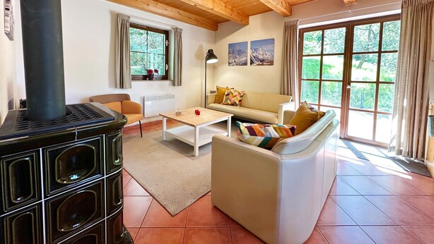 A cozy living room with a wooden ceiling, a black stove, a sofa, a coffee table, and a large window with a view of greenery. - Mountains Family Retreat (Rudník)