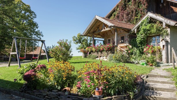 Ein rustikales Holzhaus mit einem geneigten Dach, umgeben von einem gepflegten Garten mit verschiedenen Blumen und einer Schaukel.