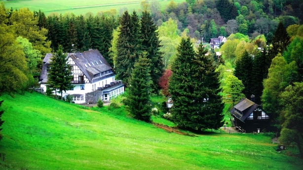 Una casa grande con un techo pronunciado, rodeada de un bosque denso y campos abiertos.