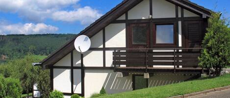 En traditionell chalet med ett sluttande tak, en satellitmottagare och utsikt över en skogbeklädd sluttning.