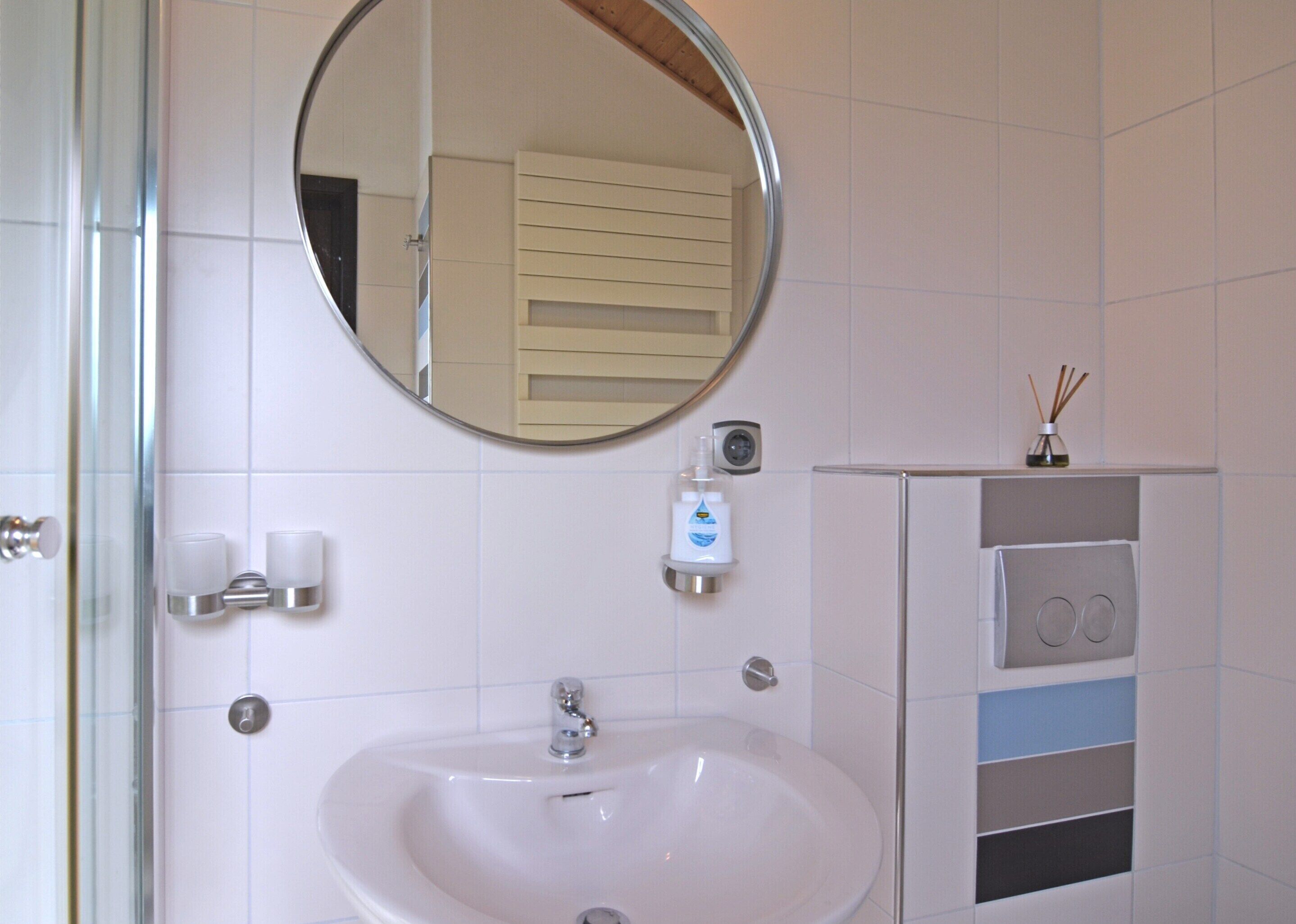 A bathroom with a white sink, a round mirror, and a small shelf with a bottle of soap and a reed diffuser.