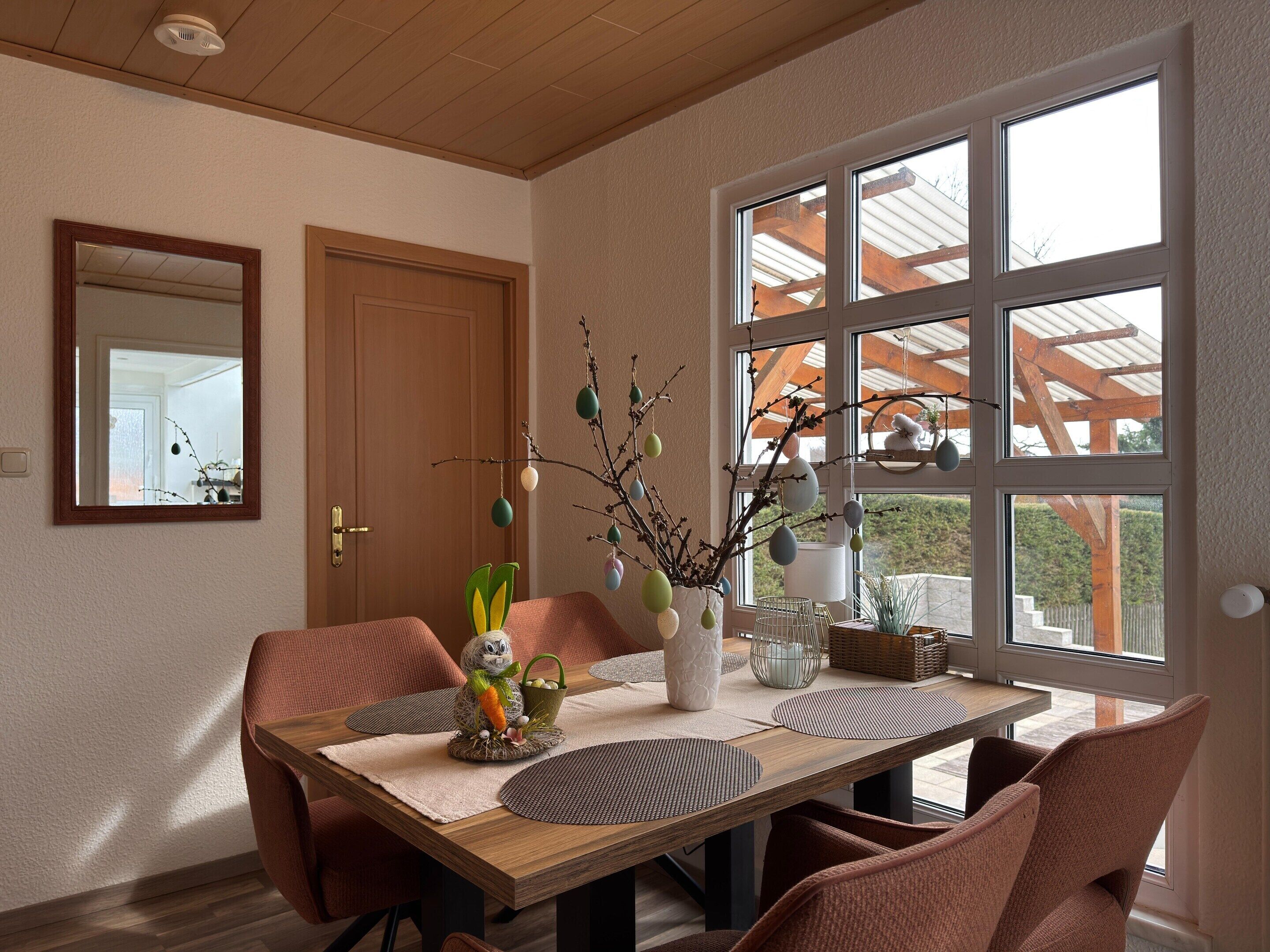 Un comedor con una mesa de madera, sillas, un florero con ramas y una ventana con vistas a una construcción de madera.