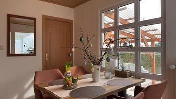 Un comedor con una mesa de madera, sillas, un florero con ramas y una ventana con vistas a una construcción de madera.