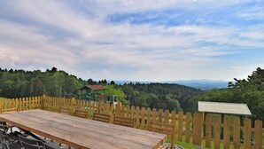 A wooden deck with a table and chairs, surrounded by a wooden fence, overlooking a forested valley with a small building in the background. - Alpine View Wellness Escape (Waldkirchen)