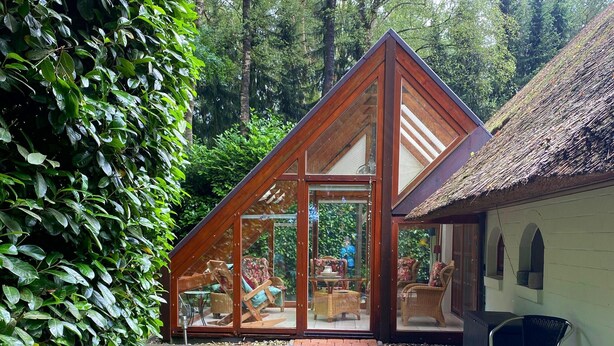 A modern house with a triangular roof, large glass windows, and a patio area with outdoor furniture. - Ferienhaus Boschhuys (Uelsen)