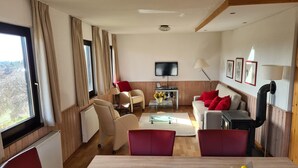 A modern living room with a wooden floor, beige sofas, a dining table with a fruit centerpiece, and a TV mounted on the wall. - Chalet in Frankenau Nahe Skipisten & Edersee (Frankenau)