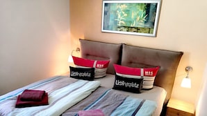 Ein ordentlich bezogenes Bett mit gestreifter Bettwäsche, zwei Kissen mit der Aufschrift 'HIS' und ein kleiner Nachttisch mit einer Lampe.