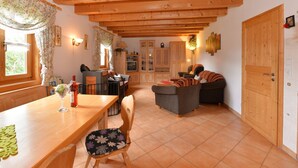 Uma sala de estar aconchegante com vigas de madeira, uma área de jantar com mesa e cadeiras, e uma televisão ao fundo.