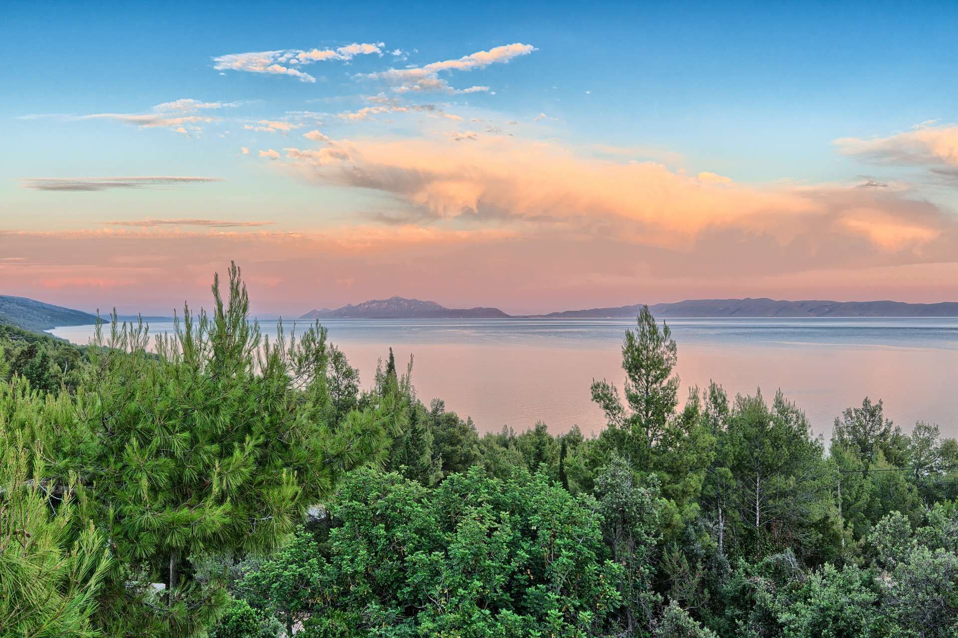 A coastal landscape with dense greenery in the foreground and a calm sea under a sunset sky.