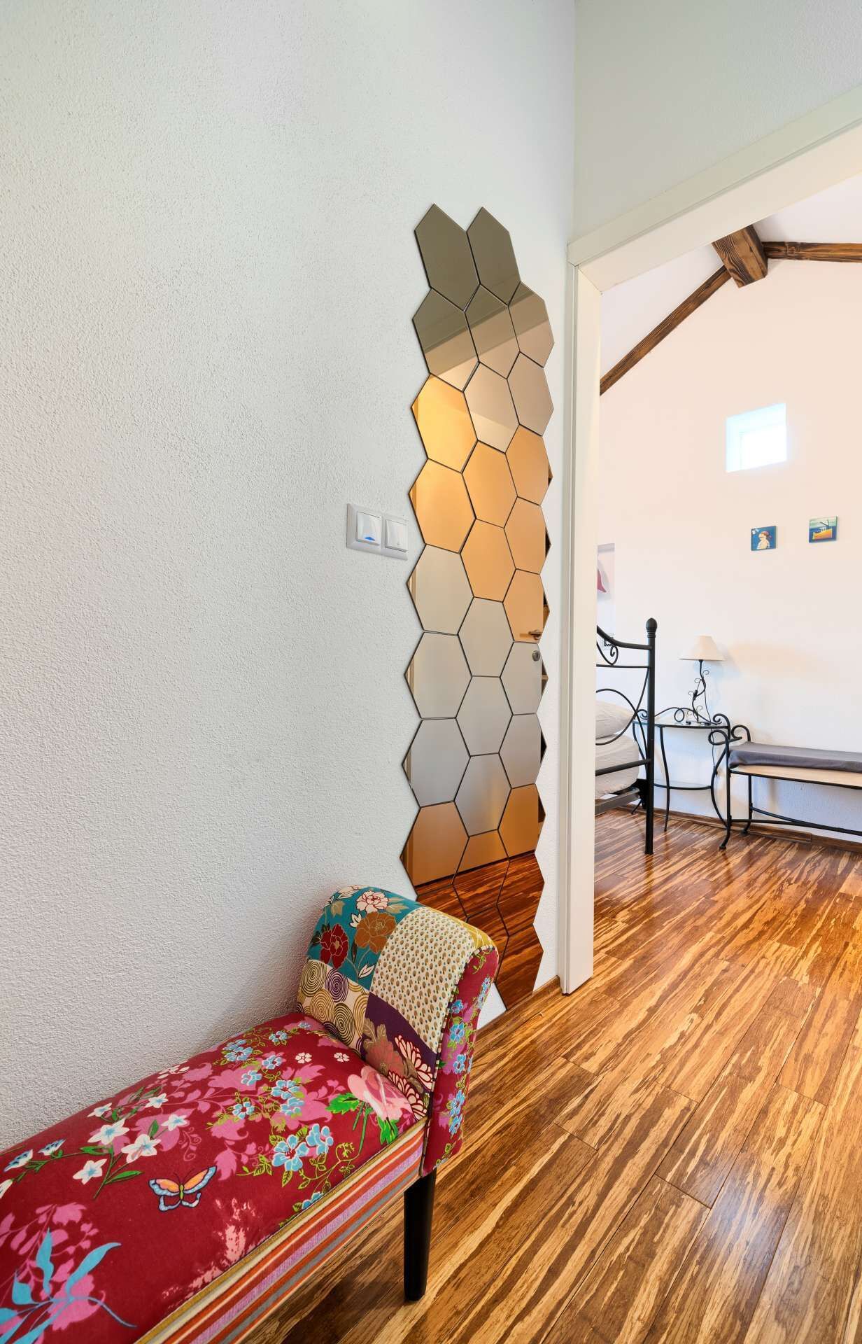 Une pièce dotée d’un plancher en bois, d’un lit recouvert d’une couvre-lit à motifs floraux colorés, d’un miroir hexagonal fixé au mur et d’une petite table avec une lampe.