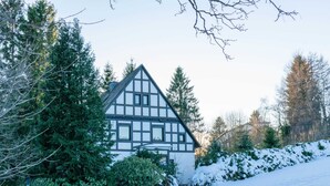 A traditional half-timbered house with a steeply pitched roof, surrounded by snow-covered ground and dense evergreen trees. - Quaint Holiday Home near Ski Area in Neuastenberg (Winterberg)