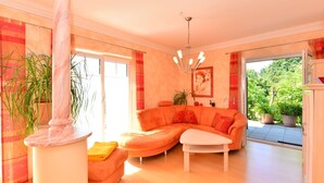 A spacious living room with a large sectional sofa, a white coffee table, and a marble column. There is a large window providing natural light and a view of greenery outside. - Apartment in Bavaria With Scenic Garden Views (Deggendorf)