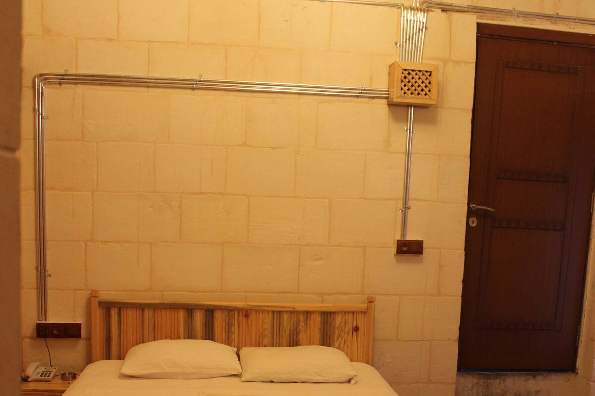 Un lit simple avec une tête de lit en bois, une couverture blanche et un plaid marron. Il y a une porte en bois sur la droite et un luminaire fixé au mur au-dessus du lit.