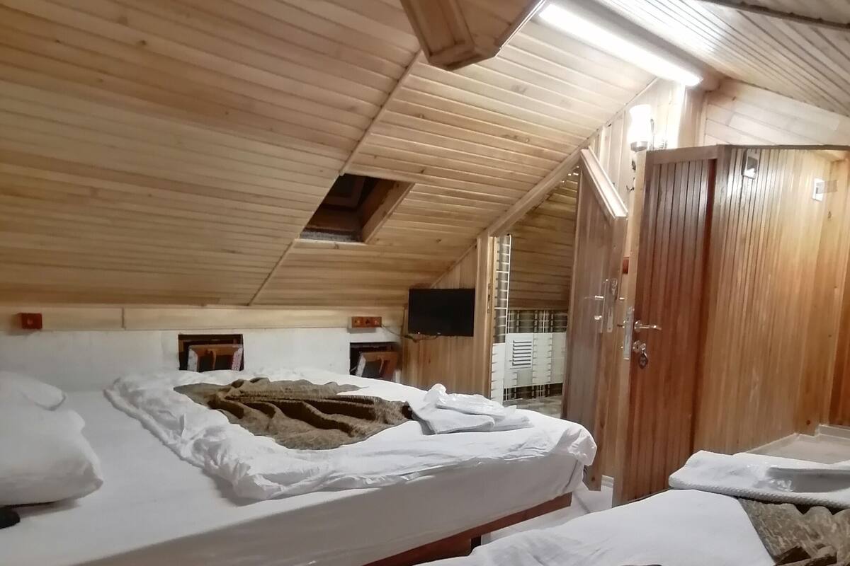Une pièce avec un plafond à lattes de bois et une hauteur sous plafond importante. On y trouve des lits avec du linge de lit blanc et une couverture marron. On peut également apercevoir une armoire en bois et un téléviseur.