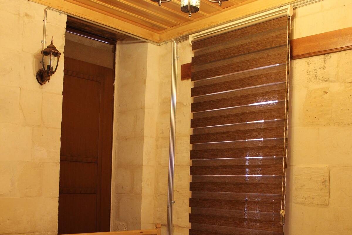 Une chambre avec un lit, une tête de lit en bois, un luminaire fixé au mur et une fenêtre avec des stores.
