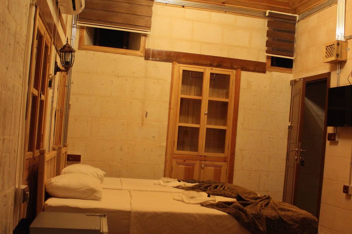 Une petite pièce chaleureuse aux murs en bois et au plafond en pente. Elle comprend deux lits, une armoire en bois et un téléviseur fixé au mur.