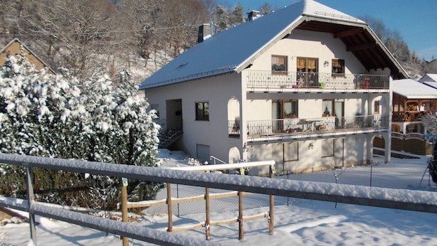 Un chalé de dos pisos con un balcón, rodeado de árboles cubiertos de nieve y una cerca de madera.