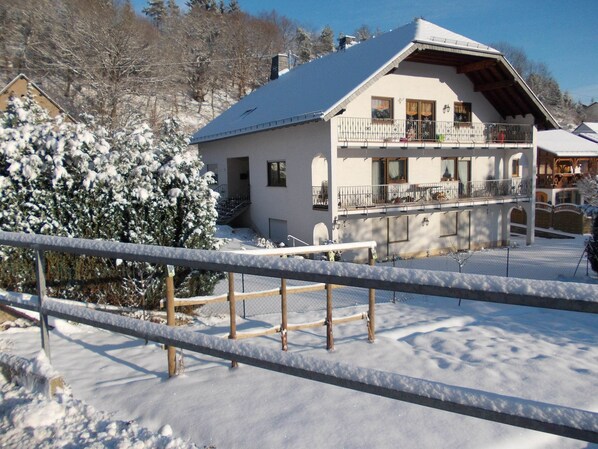 A two-story chalet with a balcony, surrounded by snow-covered trees and a wooden fence.