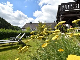 En trädgård med gula blommor, en träveranda och en byggnad med ett sluttande tak i bakgrunden.