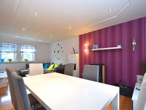 A modern living room with a white dining table, grey chairs, a grey sofa with colorful cushions, a large wall clock, and a pink accent wall. - Deifeld Countryside Escape (Medebach)