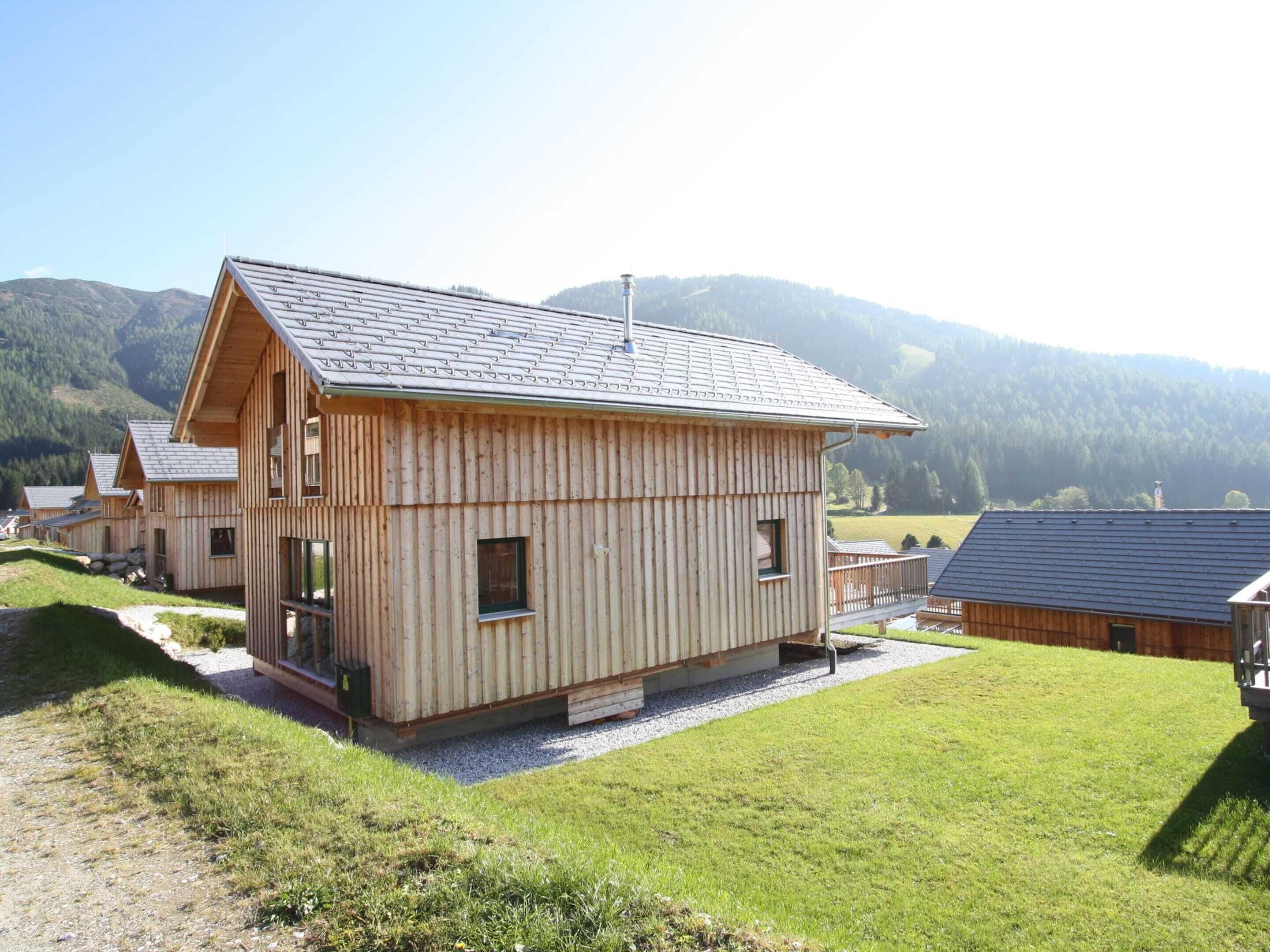 A wooden cabin with a sloped roof, surrounded by a grassy area and mountains in the background.