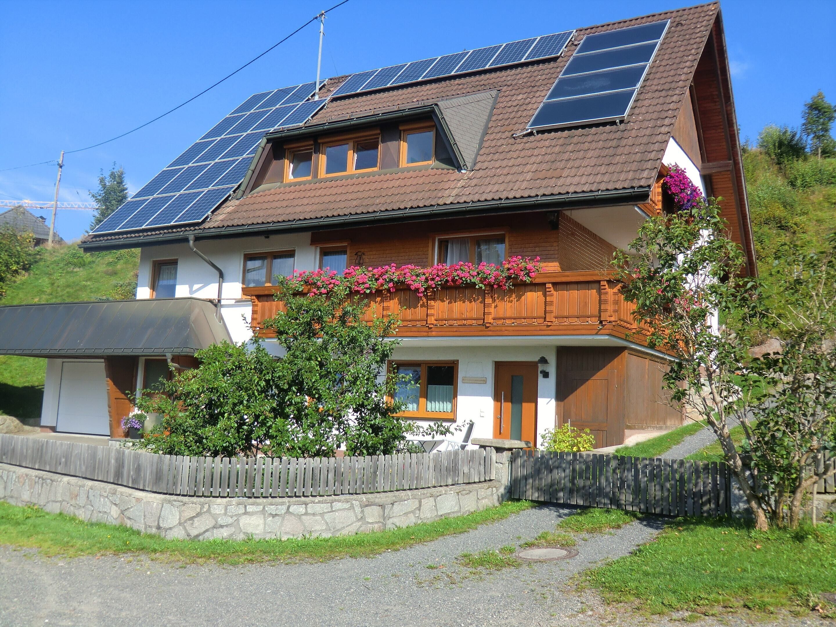 A two-story house with a sloped roof, solar panels, a wooden balcony with flowers, and a stone wall.
