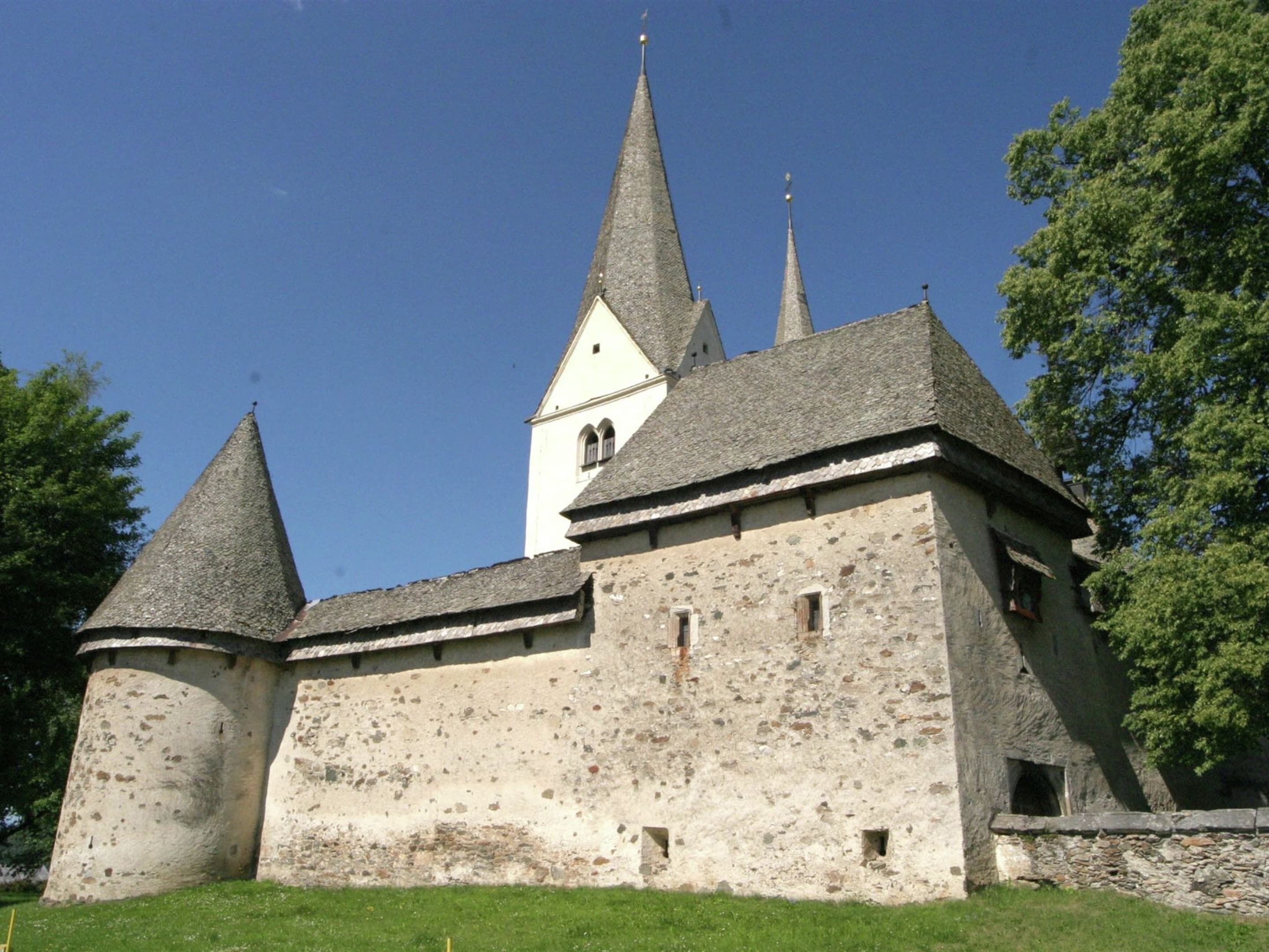 Ein historisches Gebäude mit einem zentralen Turm und zwei kleineren Türmen, umgeben von Grünflächen.