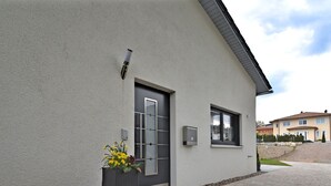 Un edificio moderno con fachada gris, una puerta oscura y una ventana. Hay una jardinera con flores amarillas frente a la puerta.