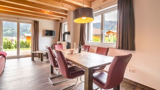 A dining area with a wooden table, red chairs, a hanging light fixture, and a TV mounted on the wall.