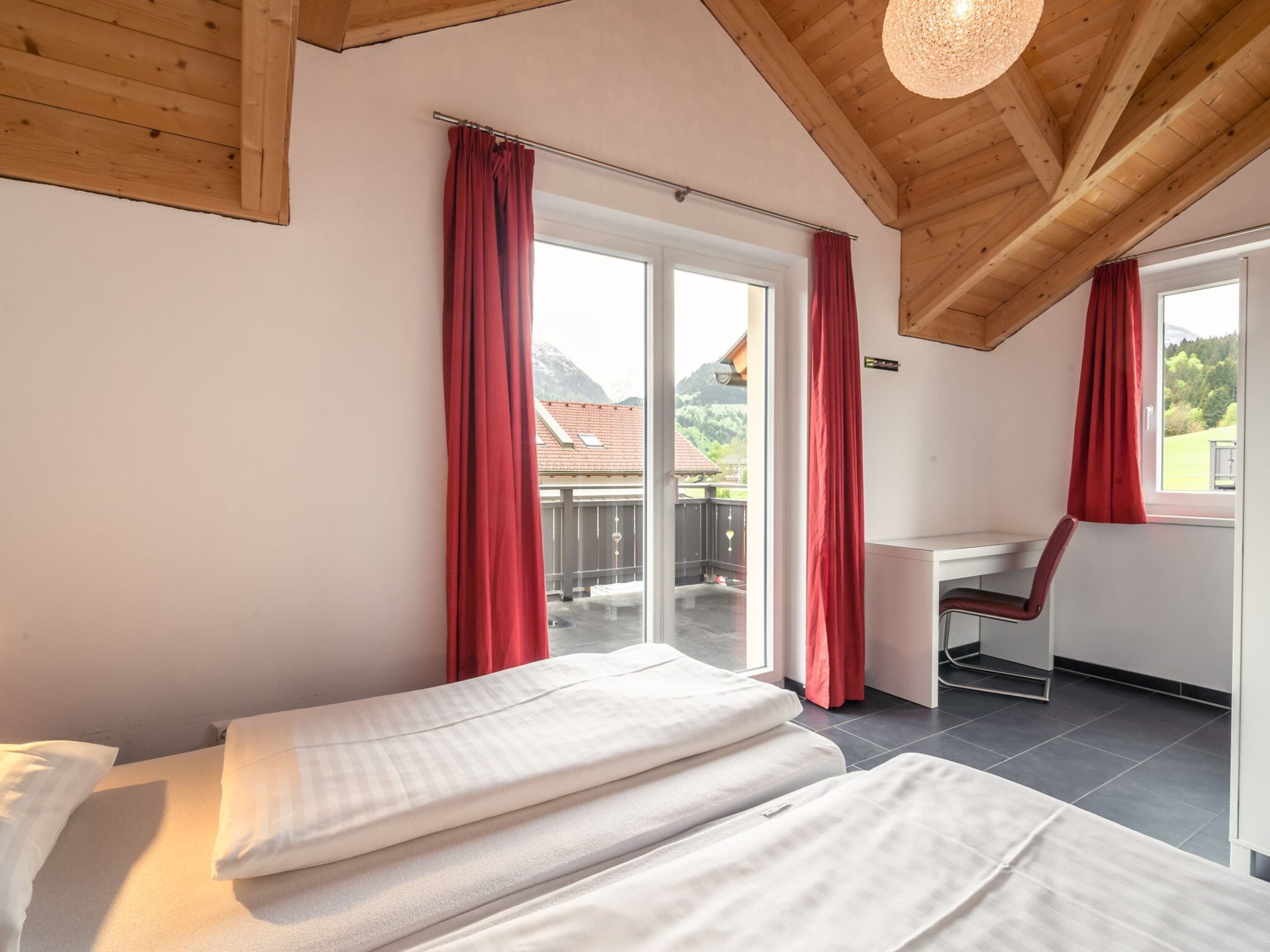A bedroom with a bed, a desk, and a chair, with a view of a balcony and mountains.